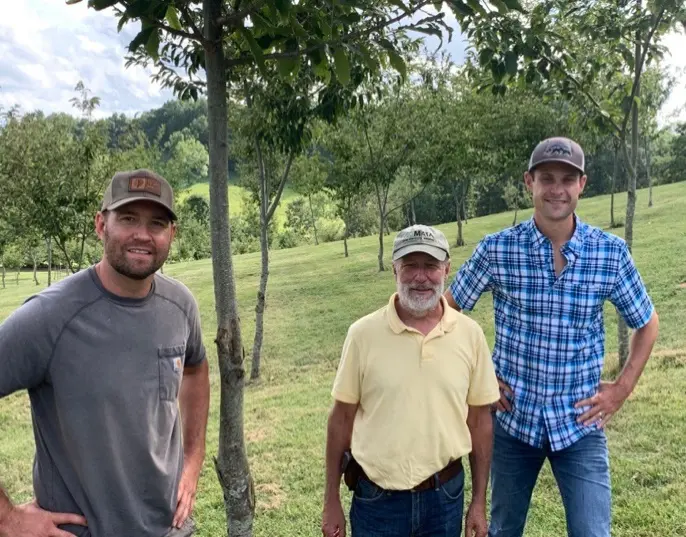 Three men standing in a field near trees.